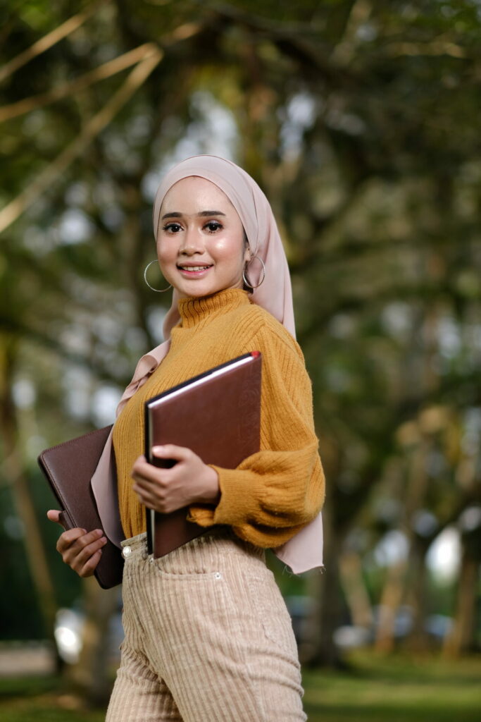 portrait smiling teenage girl wearing headscarf holding books while standing against trees